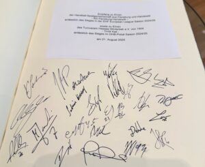 Ein Stück Papier mit deutschem Text über den THW Kiel Handball-Bundesliga Saison 2024/2025 Empfang der Landesregierung Schleswig-Holstein, liegend auf einer Seite mit zahlreichen Unterschriften in schwarzer Tinte.