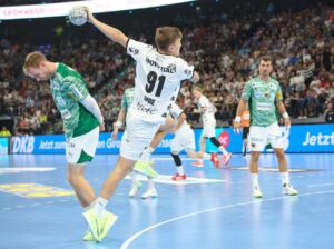 Ein Handballspieler in einem weißen Trikot springt, um den Ball zu werfen, während ein Verteidiger in einem grünen Trikot versucht, ihn zu blockieren. Andere Spieler und eine große Zuschauermenge verfolgen das Geschehen in einer hell erleuchteten Halle.