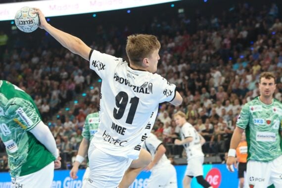 Ein Handballspieler in einem weißen Trikot springt, um den Ball zu werfen, während ein Verteidiger in einem grünen Trikot versucht, ihn zu blockieren. Andere Spieler und eine große Zuschauermenge verfolgen das Geschehen in einer hell erleuchteten Halle.