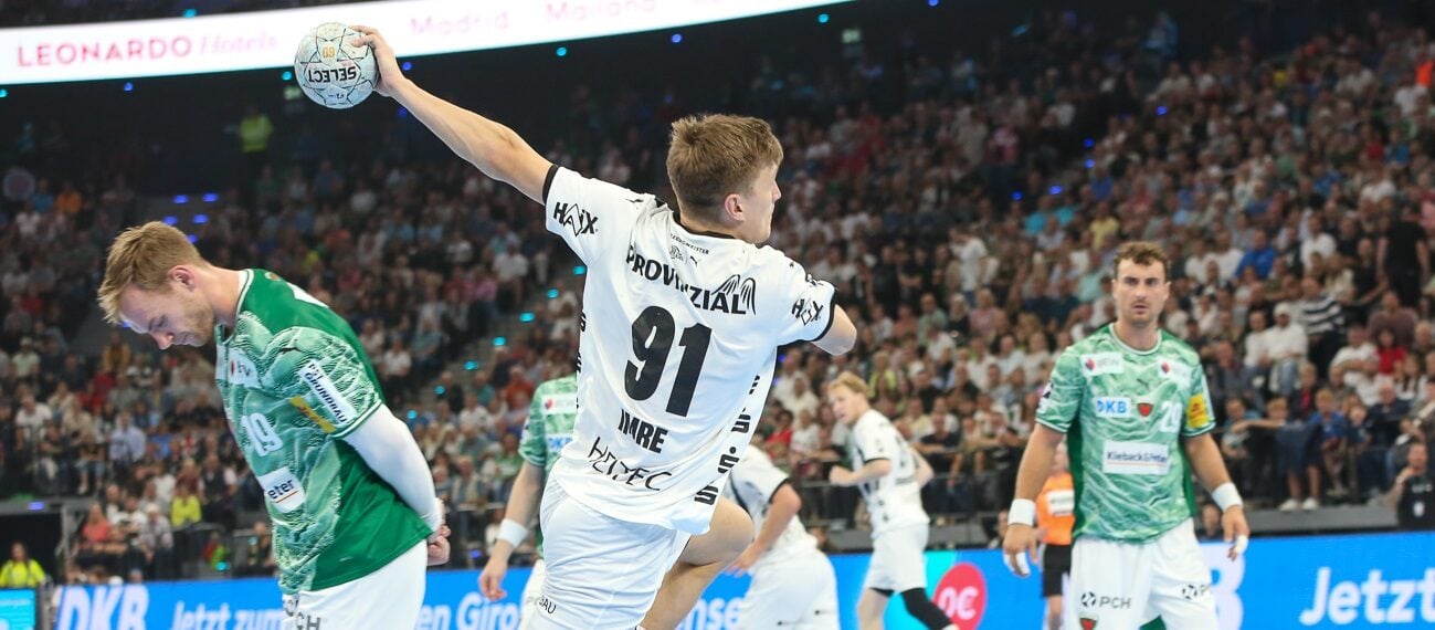 Ein Handballspieler in einem weißen Trikot springt, um den Ball zu werfen, während ein Verteidiger in einem grünen Trikot versucht, ihn zu blockieren. Andere Spieler und eine große Zuschauermenge verfolgen das Geschehen in einer hell erleuchteten Halle.