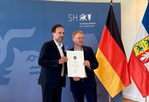 Zwei Männer in Anzügen stehen zusammen und halten eine Urkunde vor einer deutschen und einer schleswig-holsteinischen Flagge, vor einem blauen Hintergrund mit dem Schleswig-Holstein-Logo und Text.
