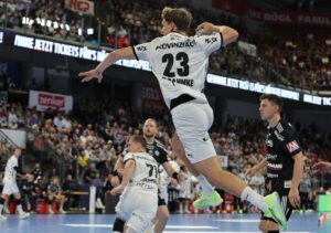 Ein Handballspieler, der ein weißes Trikot mit der Nummer 23 trägt, springt in der Luft, um den Ball zu schießen, während seine Gegner in schwarzen Trikots zusehen. Das Spiel findet in einer mit Zuschauern gefüllten Halle statt.