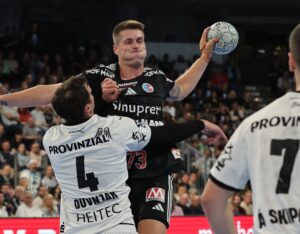 Ein Handballspieler in einem schwarzen Trikot springt zum Schuss, während er von einem Gegner in einem weißen Trikot eng verteidigt wird. Ein weiterer Spieler in Weiß ist teilweise sichtbar. Das Spiel findet in einer Halle mit Zuschauern statt.