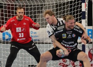 Drei männliche Handballspieler treten in der Nähe des Tores gegeneinander an. Ein Spieler in einem roten Trikot (der Torwart) ist konzentriert, während zwei Spieler in schwarz-weißen Trikots um die Kontrolle des Balls vor dem Netz kämpfen.