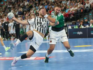 Zwei männliche Handballspieler in Aktion auf einem Spielfeld; einer in einem schwarz-weiß gestreiften Trikot springt mit dem Ball, während ein Spieler in einem grünen Trikot den Ball aggressiv abwehrt, während im Hintergrund eine Menschenmenge zusieht.