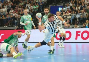 Ein Handballspieler in einem weiß-schwarzen Trikot springt mit dem Ball durch die Luft und versucht, ein Tor zu erzielen, während Spieler in grün-weißen Trikots verteidigen. Das Spiel findet in einer Halle statt, die von einer Menschenmenge beobachtet wird.
