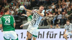 Ein Handballspieler in einem weiß-schwarz gestreiften Trikot springt und bereitet sich auf den Wurf vor, während Gegner in grünen Trikots versuchen, ihn zu blockieren. Eine Menschenmenge beobachtet das Geschehen im Hintergrund.