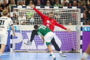 Ein Handballtorwart im roten Trikot springt mit ausgestreckten Armen, um einen Schuss abzuwehren, während ein Spieler im grünen Trikot springt, um den Ball in Richtung Tor zu werfen; im Hintergrund sind andere Spieler und Zuschauer zu sehen.