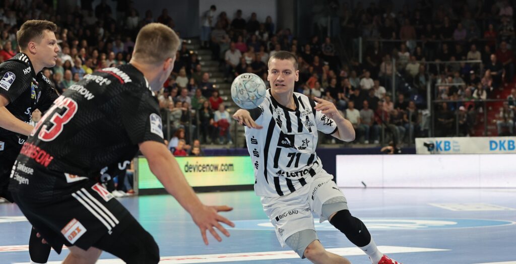 HC Erlangen - THW Kiel Ein Handballspieler in einem weiß-schwarzen Trikot wirft den Ball, während zwei Gegner in schwarzen Trikots den Ball in einer mit Zuschauern gefüllten Halle verteidigen.