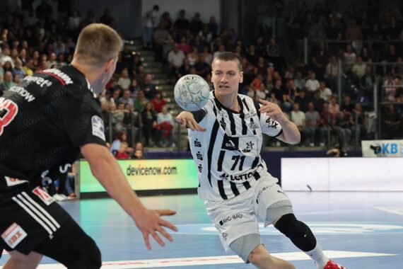 Ein Handballspieler in einem weiß-schwarzen Trikot wirft den Ball, während zwei Gegner in schwarzen Trikots den Ball in einer mit Zuschauern gefüllten Halle verteidigen.
