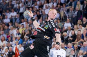 Ein Handballspieler in einem schwarzen Trikot springt und bereitet sich darauf vor, den Ball während eines Spiels zu werfen, während im Hintergrund eine Zuschauermenge zusieht.