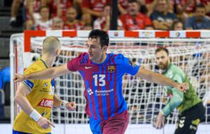 Ein Handballspieler, der ein rot-blaues Trikot mit der Nummer 13 trägt, jubelt mit ausgestreckten Armen, während im Hintergrund in der Nähe des Tores andere Spieler und ein Torwart zu sehen sind.