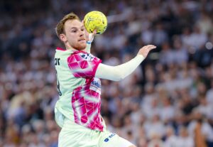 Ein männlicher Handballspieler in einem grün-rosafarbenen Trikot springt in der Luft und bereitet sich auf den Wurf eines gelben Handballs vor. Im Hintergrund ist eine verschwommene Menschenmenge zu sehen, die eine intensive Spielatmosphäre suggeriert.
