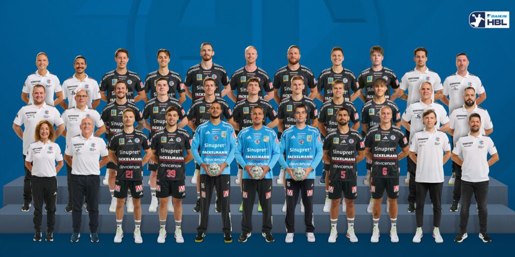 26_HCE_Teambild Eine professionelle Männerhandballmannschaft posiert für ein Gruppenfoto in Trikots, flankiert von Trainern und Betreuern in weißen Oberteilen, die alle in Reihen vor einem blauen Hintergrund mit einem Liga-Logo in der Ecke stehen und sitzen.