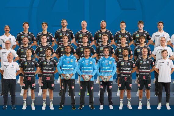 Eine professionelle Männerhandballmannschaft posiert für ein Gruppenfoto in Trikots, flankiert von Trainern und Betreuern in weißen Oberteilen, die alle in Reihen vor einem blauen Hintergrund mit einem Liga-Logo in der Ecke stehen und sitzen.