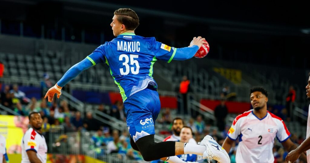 26_PM_Domen_Titel Ein männlicher Handballspieler in einem blauen Trikot mit der Nummer 35 springt in der Luft, um den Ball zu werfen, während seine Gegner in weißen und roten Trikots in einer Halle mit Zuschauern im Hintergrund zusehen.