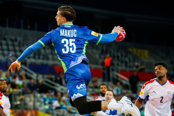 Ein männlicher Handballspieler in einem blauen Trikot mit der Nummer 35 springt in der Luft, um den Ball zu werfen, während seine Gegner in weißen und roten Trikots in einer Halle mit Zuschauern im Hintergrund zusehen.