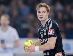 Ein männlicher Handballspieler in einem schwarzen Trikot hält einen gelben Handball und schaut konzentriert während eines Spiels. Der Hintergrund ist unscharf, ein anderer Spieler ist zu sehen.