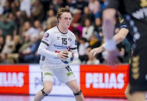 Ein männlicher Handballspieler in einem weißen Trikot, der einen Ball hält, bereitet sich auf einen Spielzug vor, während im Hintergrund Zuschauer und eine digitale Anzeigetafel zu sehen sind.