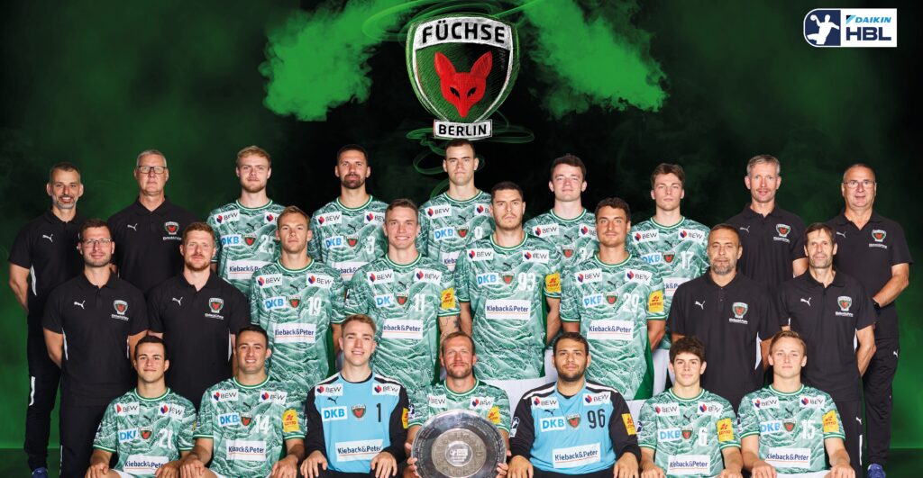 Mannschaftsfoto des Handballvereins Füchse Berlin; Spieler und Betreuer posieren in grünen Trikots mit Sponsorenlogos, hinter einem großen Mannschaftslogo und grünem Raucheffekt. Eine Person im Vordergrund hält eine schildförmige Trophäe.