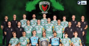 Mannschaftsfoto des Handballvereins Füchse Berlin; Spieler und Betreuer posieren in grünen Trikots mit Sponsorenlogos, hinter einem großen Mannschaftslogo und grünem Raucheffekt. Eine Person im Vordergrund hält eine schildförmige Trophäe.