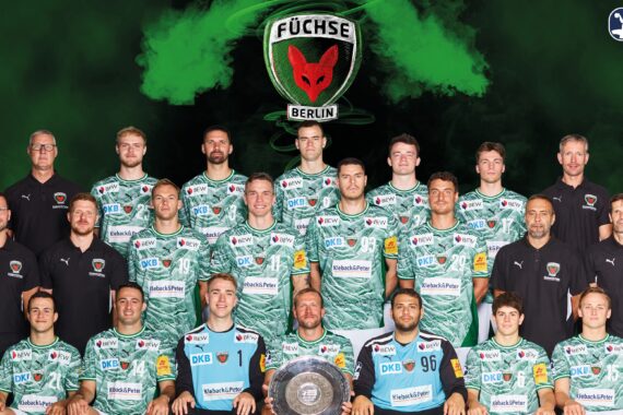 Mannschaftsfoto des Handballvereins Füchse Berlin; Spieler und Betreuer posieren in grünen Trikots mit Sponsorenlogos, hinter einem großen Mannschaftslogo und grünem Raucheffekt. Eine Person im Vordergrund hält eine schildförmige Trophäe.