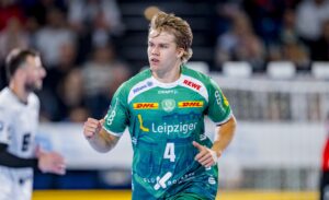 Ein männlicher Handballspieler in einem grünen Trikot mit der Nummer 4, der während eines Spiels läuft. Der Hintergrund ist unscharf, ein anderer Spieler und ein Tor sind zu sehen. Auf dem Trikot sind verschiedene Sponsorenlogos zu sehen.