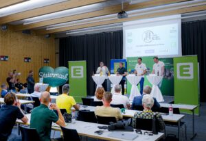 Ein Gremium von vier Rednern steht an Stehtischen auf einer Bühne vor einem Publikum, mit grünen Bannern und einem großen Ballonherz mit der Aufschrift "Steiermark" in einem modernen Konferenzraum. Ein Fotograf hält die Veranstaltung fest.