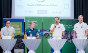 Vier Männer stehen an hohen runden Tischen und nehmen an einer Podiumsdiskussion teil. Ein Mann spricht in ein Mikrofon. Hinter ihnen hängen Banner und Schilder mit Veranstaltungsinformationen und Sponsorenlogos in deutscher Sprache. Auf jedem Tisch stehen Flaschen mit Getränken.