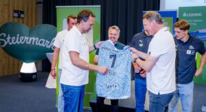 Eine Gruppe von Männern in Markenshirts steht zusammen, während ein Mann ein blau-weißes Fußballtrikot mit der Nummer 7 und dem Namen DIDI hochhält. Im Hintergrund ist ein großes herzförmiges Schild mit der Aufschrift "Steiermark" zu sehen.