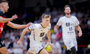 Ein männlicher Handballspieler in einem weißen Trikot läuft mit einem gelben Ball, während ein Gegner in einem rot-schwarzen Trikot nach ihm greift. Ein weiterer weiß gekleideter Spieler ist im Hintergrund zu sehen. Die Szene findet in einer Halle während eines Spiels statt.