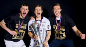 Drei begeisterte männliche Athleten feiern, lächeln und jubeln, tragen Medaillen und halten einen großen Pokal. Zwei von ihnen tragen schwarze "EHF EUROPEAN LEAGUE WINNER"-Shirts, während der mittlere Sportler eine weiße Uniform trägt. Schwarzer Hintergrund.