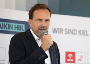Ein Mann in einem dunklen Anzug hält ein Mikrofon in der Hand und spricht auf einer Veranstaltung. Er steht vor einem Hintergrund mit dem Text WIR SIND KIEL und mehreren Logos.