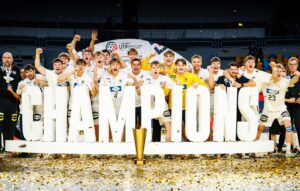 Eine jubelnde Jugendsportmannschaft in weißen Trikots feiert hinter großen CHAMPIONS-Buchstaben, mit einer Trophäe und goldenem Konfetti auf dem Boden, die Begeisterung und den Sieg in einer hell erleuchteten Arena zeigen.