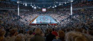 Eine überfüllte Halle mit Zuschauern, die ein professionelles Handballspiel auf einem leuchtend blauen Spielfeld verfolgen, während die Spieler in Aktion sind und die Stadionbeleuchtung die Szene beleuchtet.