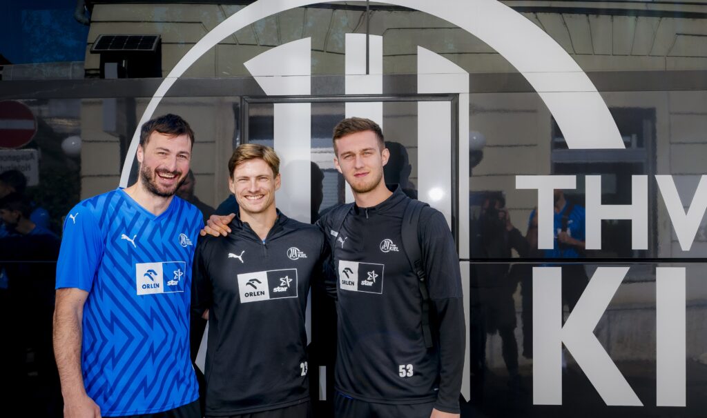 HANDBALL 2025/26: THW Kiel in der Saisonvorbereitung Drei männliche Fußballspieler stehen lächelnd vor einem Mannschaftsbus mit einem großen weißen Logo. Zwei tragen passende schwarze Trainingsanzüge, einer ein blaues Hemd. Sie posieren mit umschlungenen Armen und schauen in die Kamera.