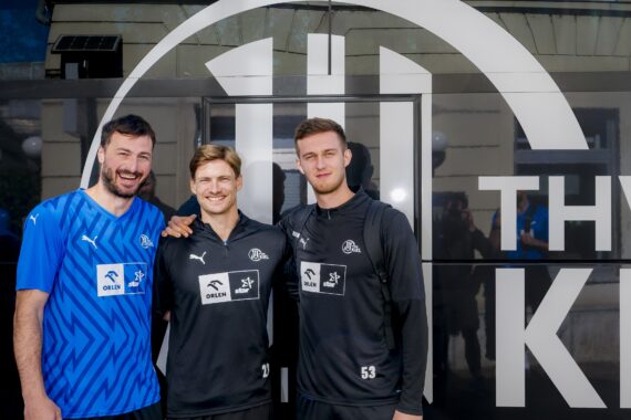 Drei männliche Fußballspieler stehen lächelnd vor einem Mannschaftsbus mit einem großen weißen Logo. Zwei tragen passende schwarze Trainingsanzüge, einer ein blaues Hemd. Sie posieren mit umschlungenen Armen und schauen in die Kamera.