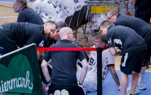 Eine Gruppe von Sportmedizinern und Trainern kümmert sich um einen verletzten männlichen Athleten, der während eines Hallenspiels auf dem Boden sitzt. Der Sportler schaut zu ihnen auf, während sie mit ihm sprechen und ihn untersuchen. Eine rote Absperrung und ein grünes Werbebanner sind zu sehen.