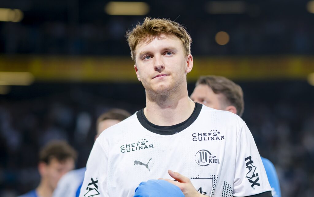 HANDBALL 2024/25: THW Kiel - ThSV Eisenach Ein männlicher Sportler mit kurzen blonden Haaren, der ein weißes Sporttrikot mit schwarzen Akzenten und Sponsorenlogos trägt, steht auf einem Spielfeld und hält einen blauen Gegenstand in der Hand; im Hintergrund sind unscharfe Menschen und Stadionlichter zu sehen.