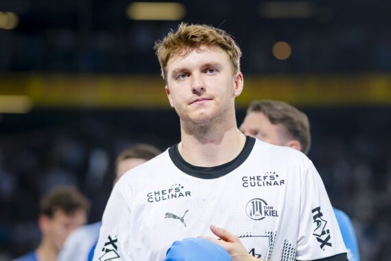 Ein männlicher Sportler mit kurzen blonden Haaren, der ein weißes Sporttrikot mit schwarzen Akzenten und Sponsorenlogos trägt, steht auf einem Spielfeld und hält einen blauen Gegenstand in der Hand; im Hintergrund sind unscharfe Menschen und Stadionlichter zu sehen.