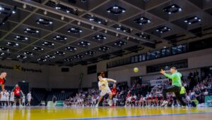 Ein Handballspieler in einem weißen Trikot springt zum Torschuss, während der Torwart in Grün sich darauf vorbereitet, den Schuss zu blocken. Die Zuschauer verfolgen das Geschehen in einer hell erleuchteten Halle.