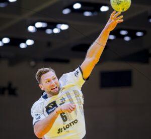 Ein männlicher Handballspieler in einem weißen Trikot, in der Luft, im Begriff, einen gelben Ball mit der rechten Hand zu werfen, konzentriert und intensiv, unter den hellen Lichtern der Arena.