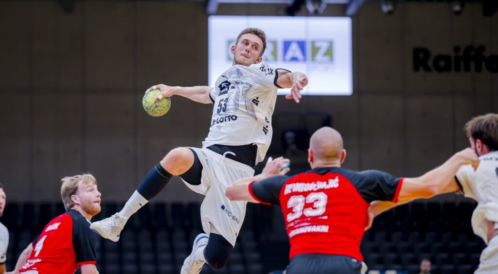 Ein Handballspieler in einem weißen Trikot springt in der Luft, um den Ball zu werfen, während zwei Verteidiger in roten und schwarzen Trikots versuchen, ihn während eines Spiels in einer Halle zu blockieren.