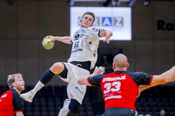 Ein Handballspieler in einem weißen Trikot springt in der Luft, um den Ball zu werfen, während zwei Verteidiger in roten und schwarzen Trikots versuchen, ihn während eines Spiels in einer Halle zu blockieren.