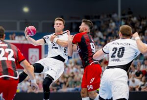 Ein Handballspieler in einem weißen Trikot springt und bereitet sich auf den Wurf vor, während er von Gegnern in roten und schwarzen Trikots während eines überfüllten Hallenspiels eng verteidigt wird.