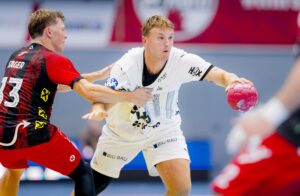 Zwei männliche Handballspieler in Aktion; der eine im weißen Trikot hält einen rosafarbenen Ball, während ein Spieler in einem rot-schwarzen Trikot versucht, ihn zu blocken. Ein weiterer Spieler in Rot ist teilweise im Vordergrund zu sehen.