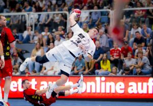 Ein Handballspieler in einem weißen Trikot springt in der Luft und bereitet sich auf den Wurf vor, während ein Verteidiger in Rot zu Boden fällt. Die Zuschauer beobachten das intensive Spiel von den überfüllten Tribünen im Hintergrund.