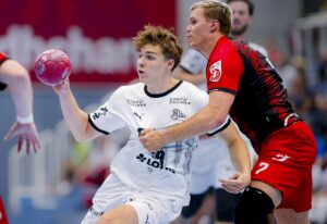 Zwei männliche Handballspieler in Aktion während der THW Kiel Handball Vorbereitung Saison 2025/2026 Sommercup Hard; der Spieler in Weiß hält den Ball und weicht einem nahen Verteidiger in Rot und Schwarz aus, während sich beide intensiv auf das Spielfeld konzentrieren.
