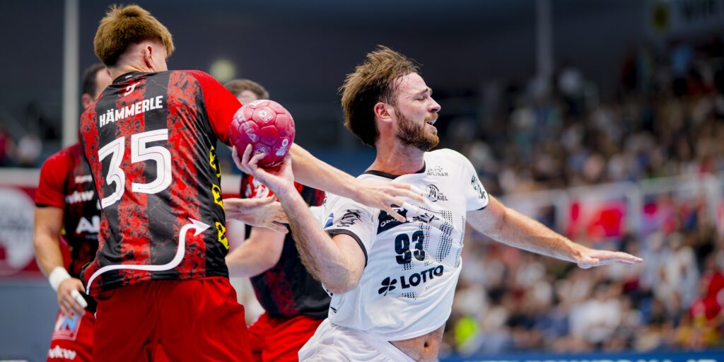 HANDBALL 2025/26: THW Kiel in der Saisonvorbereitung Ein Handballspieler in einem weißen Trikot versucht, während eines Spiels vor einer jubelnden Menge an einem Gegner in einem rot-schwarzen Trikot vorbeizukommen, der einen rosa Ball hält.