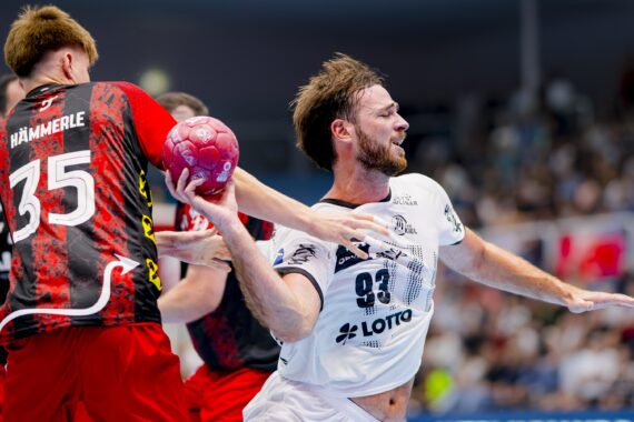Ein Handballspieler in einem weißen Trikot versucht, während eines Spiels vor einer jubelnden Menge an einem Gegner in einem rot-schwarzen Trikot vorbeizukommen, der einen rosa Ball hält.